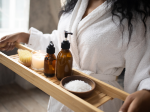 A lady in her dressing gown holding a bath rack with a candle and bath bottles setting up a spa retreat for a staycation